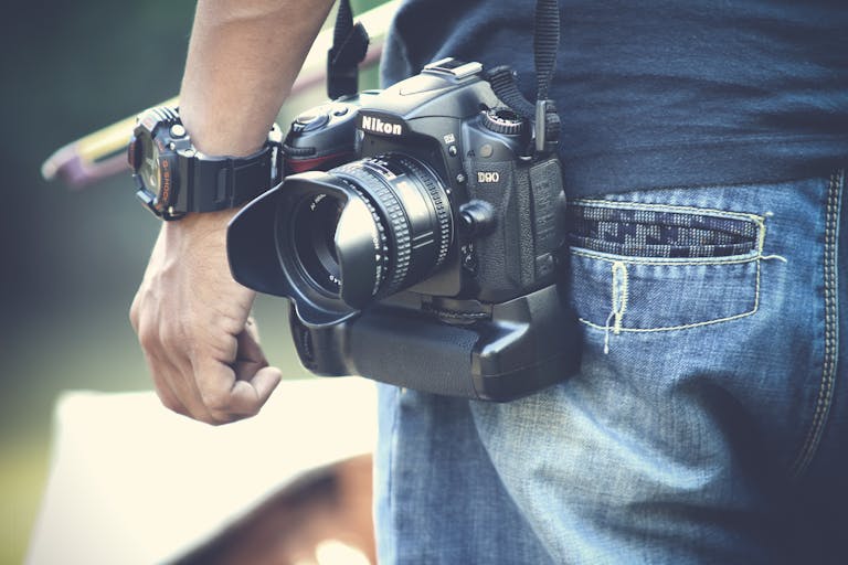 A photographer holds a DSLR camera, wearing casual denim jeans and wristwatch outdoors.