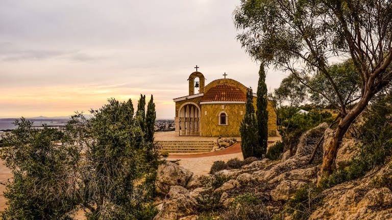 A picturesque historic church in Cyprus at sunset, surrounded by trees and rocks.