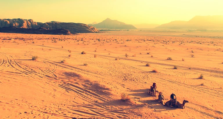 A striking warm-toned desert scene with camels and riders against majestic mountains.