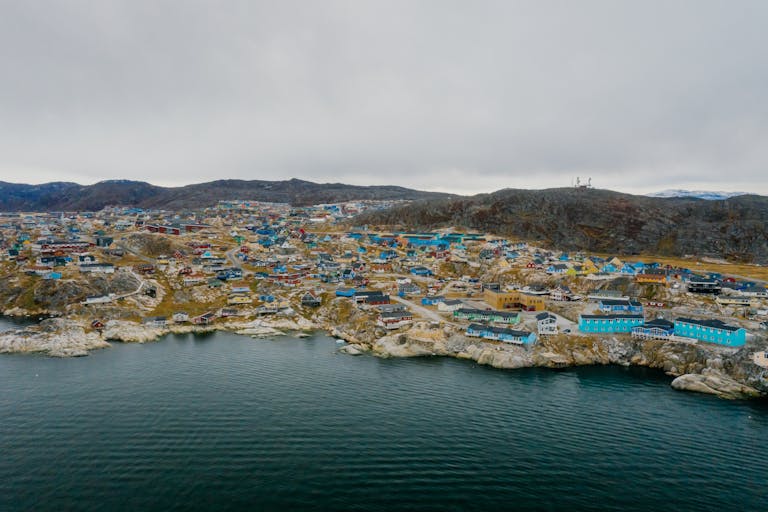 Aerial view of a colorful coastal townscape in Greenland with rugged terrain and serene waters.