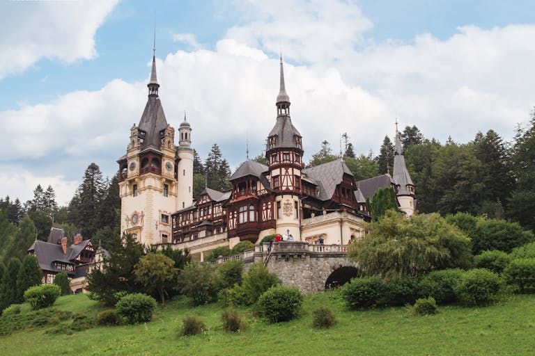 Peles Castle surrounded by lush greenery in Sinaia, Romania, under a partly cloudy sky.