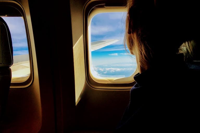 Silhouette of a woman looking out an airplane window at clouds and blue sky.