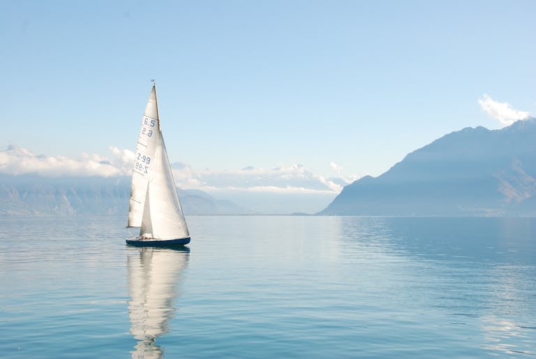 Sailing boat glides peacefully on a misty lake surrounded by mountains, under clear daylight.