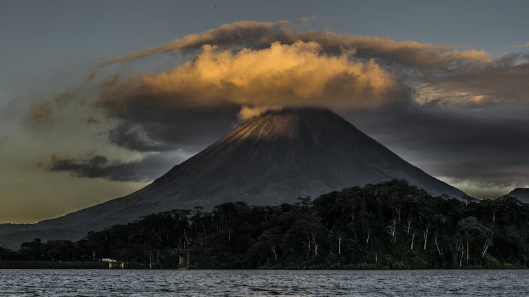 Stunning view of Arenal Volcano with glowing clouds at dusk in Costa Rica, surrounded by lush forest and tranquil waters.